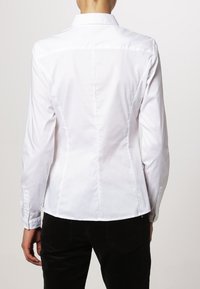 Persona vestita con una camicia bianca a maniche lunghe con colletto e pantaloni neri, vista da dietro su uno sfondo semplice.