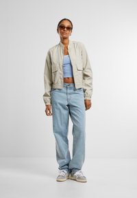 Jachetă bomber bej deschis, cu un croi scurt, dou�ă buzunare frontale, asociată cu un top albăstrui și blugi largi din denim deschis.