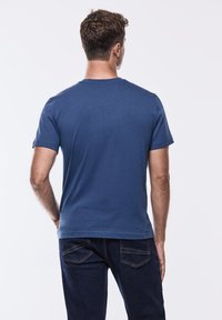 Modré pánské bavlněné tričko s krátkým rukávem, kulatým výstřihem a hladkým povrchem. Na sobě má tmavě modré džíny, pohled zezadu.
