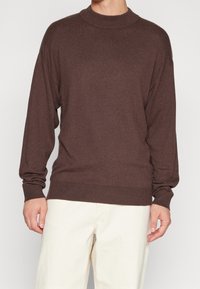 Brązowy sweter z wysokim kołnierzem, długimi rękawami i prążkowanymi mankietami. Wykonany z miękkiego materiału. W połączeniu z kremowymi spodniami.