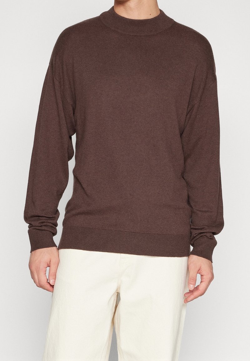 Brązowy sweter z wysokim kołnierzem, długimi rękawami i prążkowanymi mankietami. Wykonany z miękkiego materiału. W połączeniu z kremowymi spodniami.