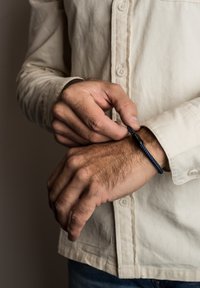 Braccialetto intrecciato blu con un fermaglio nero, indossato su un polso, abbinato a una camicia a bottoni chiara. Dettaglio visibile della mano.