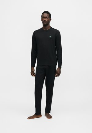 Mann steht barfuß und trägt ein schwarzes Langarmshirt mit kleinem grünem Krokodil-Logo und passende schwarze Hose auf schlicht weißem Hintergrund.