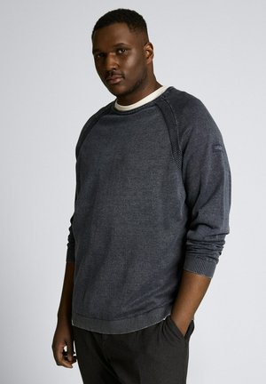 Schwarzer Mann trägt einen dunkelgrauen Pullover mit langen Ärmeln und schwarze Hose, steht mit einer Hand in der Hosentasche vor einem schlichten hellen Hintergrund.