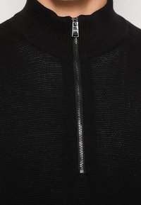 Pull en maille noire avec une fermeture éclair métallique à mi-hauteur fermée jusqu'au col, montré sur la partie supérieure de la poitrine et du cou d'une personne.