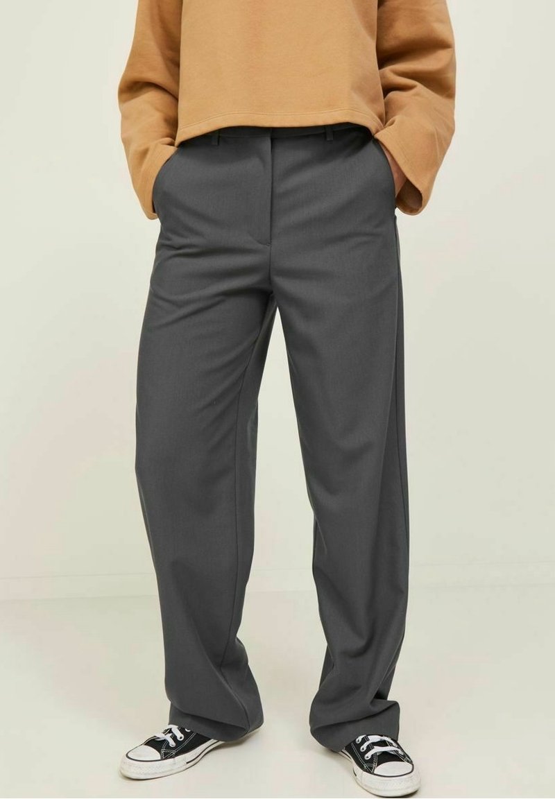 Personne portant un pantalon ample gris foncé, un haut à manches longues beige et des baskets noires et blanches, les mains dans les poches.
