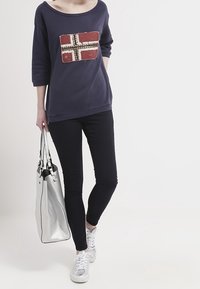 Felpa blu navy con design della bandiera rosso e bianco a rilievo, abbinata a jeans skinny neri e sneaker argento. Tiene in mano una borsa tote argento.