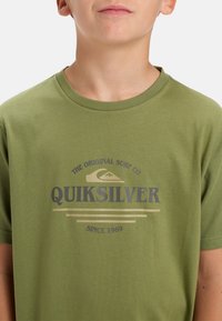 Olivově zelené tričko vyrobené z měkké bavlny. Obsahuje logo Quiksilver v tmavě šedé barvě s grafickým znázorněním vlny a textem "THE ORIGINAL SURF CO" nad ním.