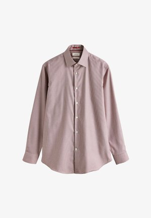 Mann mit grauem Bart, der ein rosa Hemd, eine gemusterte Krawatte, graue Anzughose und schwarze Slipper trägt, steht in einem Raum mit weißen Paneelwänden und Holzboden.