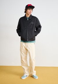 Veste zippée noire avec col, pantalon crème et baskets bleu clair. La veste présente un petit logo blanc sur la poitrine gauche.