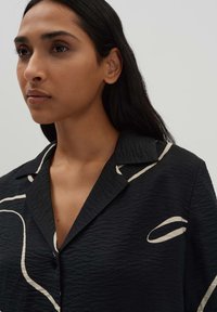 Chemise noire texturée avec un col en V, ornée de lignes abstraites blanches et de boutons sur le devant, vue sur la partie supérieure du corps d'un modèle.