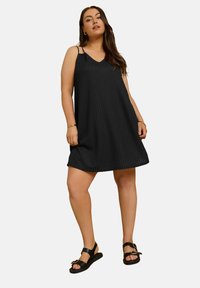 Musta ilmanhiha dressi, jossa on V-pääntie ja teksturoitu, uurrettu kuvio. Mallilla on päällään mustat sandaalit, joissa on soljendetail. 