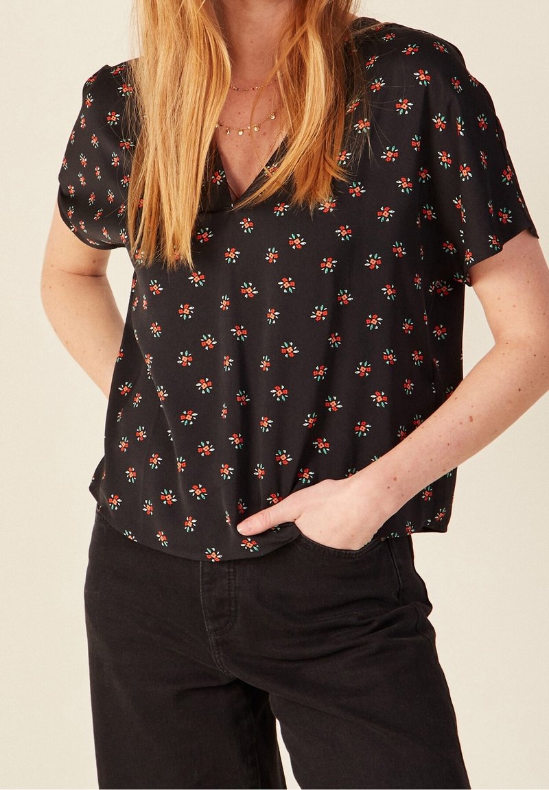 Femme portant un chemisier noir à manches courtes avec un petit imprimé floral rouge et blanc, associé à un pantalon noir, main dans la poche.