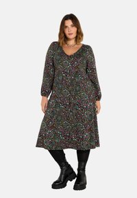 Dlouhý rukáv zavinovací šaty z tmavé látky s barevnými paisley vzory, s výstřihem do V a splývavým A-liniovým siluetem. V kombinaci s robustními černými botami.