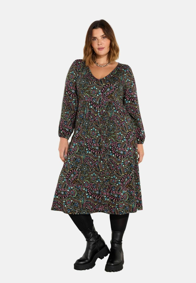 Dlouhý rukáv zavinovací šaty z tmavé látky s barevnými paisley vzory, s výstřihem do V a splývavým A-liniovým siluetem. V kombinaci s robustními černými botami.