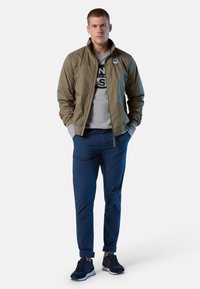 Giubbotto bomber verde oliva con colletto alto, felpa grigia sotto, pantaloni navy e scarpe sportive scure con lacci.