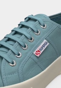 Gros plan sur une basket en toile bleu sarcelle avec des lacets de la même couleur passant à travers des œillets en métal et une étiquette de marque Superga blanche sur le côté.