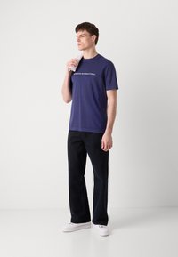 Tricou din bumbac bleumarin cu mânecă scurtă, cu text alb "Barbour International". Asortat cu pantaloni negri și adidași albi.