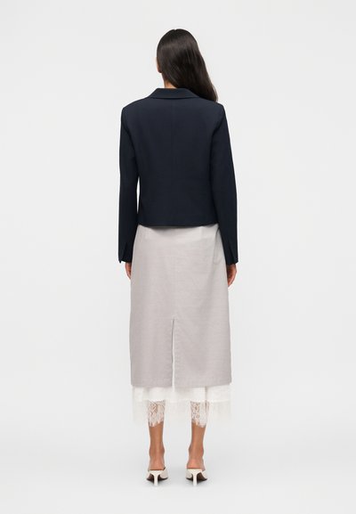Γυναίκα που στέκεται με την πλάτη να βλέπει την κάμερα, φορώντας μαύρο κοντό σακάκι, ανοιχτό γκρι φούστα με κεντρική σχισμή και λευκό εσωτερικό στρώμα με δαντέλα, και ψηλοτάκουνα παπούτσια.