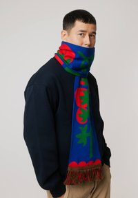 Wełniany szalik z niebieskim tłem, ozdobiony żywymi czerwonymi kwiatowymi wzorami i zielonymi akcentami, wykończony frędzlami.