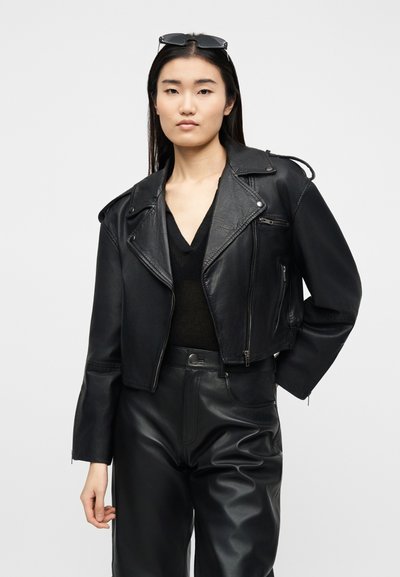 Νεαρή γυναίκα που φοράει μαύρο κοντό δερμάτινο σακάκι, μαύρο μπλουζάκι με V λαιμόκοψη, ταιριαστό μαύρο δερμάτινο παντελόνι και γυαλιά ηλίου πάνω στο κεφάλι της, μπροστά σε λευκό φόντο.