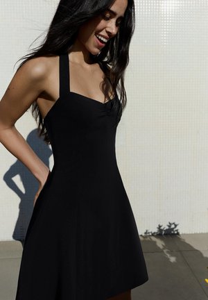 Woman smiling and looking down wearing a sleeveless black dress with wide straps, standing against a tiled light wall in sunlight.