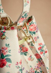 Blomstrede stofhåndtaske med en struktureret hvid baggrund, prydet med pink, røde og grønne blomster. Gyldent hardware og en grøn accentrem.