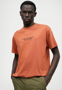 Tricou cu mâneci scurte, gât rotund, de culoare portocalie arsă, realizat din bumbac, având un text logo imprimat în negru în centrul său. Purtați cu pantaloni verzi.