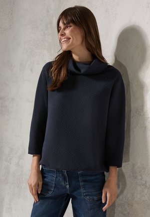Cowl-Neck-Pullover in Dunkelblau mit strukturiertem geometrischem Muster, kombiniert mit Jeans. Verfügt über Dreiviertelärmel und einen lässigen Schnitt.