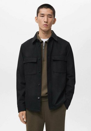 Black button-up jacket with two chest pockets, made of a textured fabric. Worn over an olive green zip-neck top.