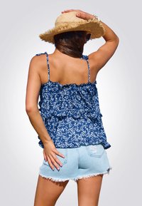 Top de tirantes azul con estampado floral y volantes, combinado con pantalones cortos de mezclilla desgastados. La tela es ligera; las correas son ajustables. El fondo es neutro.