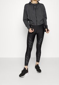 Svart kort hoodie med dragkedja framtill, kombinerad med högmidjade svarta leggings med en strukturerad mörk mönstring. Svarta träningsskor.