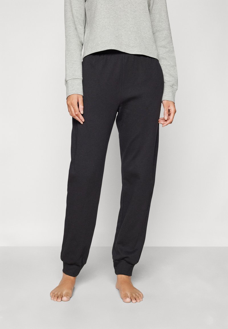 Persoon in zwarte joggingbroek en een lichtgrijs shirt met lange mouwen, staat blootsvoets op een lichtgrijze vloer tegen een plain muur.