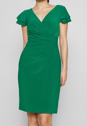 Femme portant une robe verte longueur genou avec des manches courtes à volants et un décolleté en V, présentant des détails froncés sur le devant.