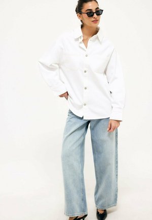 Woman wearing black sunglasses, a white button-up shirt, light blue wide-leg jeans, and black heels, standing with one hand on hip.