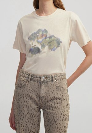 Kvinde iført en cremefarvet T-shirt med et abstrakt farvemønster og højtaljede beige jeans med mørkt plettet tryk.