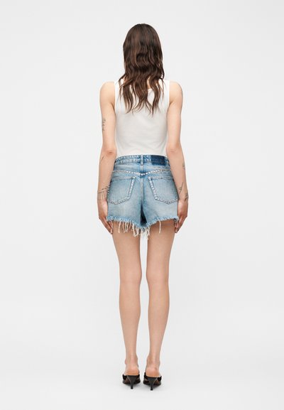 Γυναίκα με μακριά κυματιστά μαλλιά φοράει λευκό τοπ χωρίς μανίκια, ξεφτισμένα ανοιχτό μπλε τζιν σορτς και μαύρα σανδάλια με τακούνι, στέκεται μπροστά σε απλό φόντο.