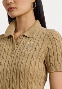 Polo en maille torsadée beige avec des manches courtes, doté d'un col côtelé et de boutons ; logo brodé sur la poitrine en blanc.
