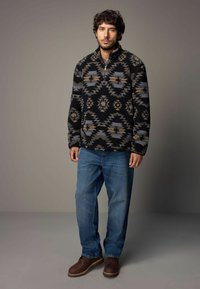 Czarny polarowy sweter z geometrycznymi wzorami w odcieniach beżu i niebieskiego, z kołnierzem na pół zamka, zestawiony z niebieskimi dżinsami i brązowymi butami.