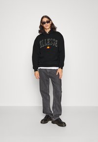 Zwarte hoodie met een frontzak en het "Ellesse" logo; gecombineerd met donkergrijze jeans en zwarte schoenen met gele stikselaccenten.