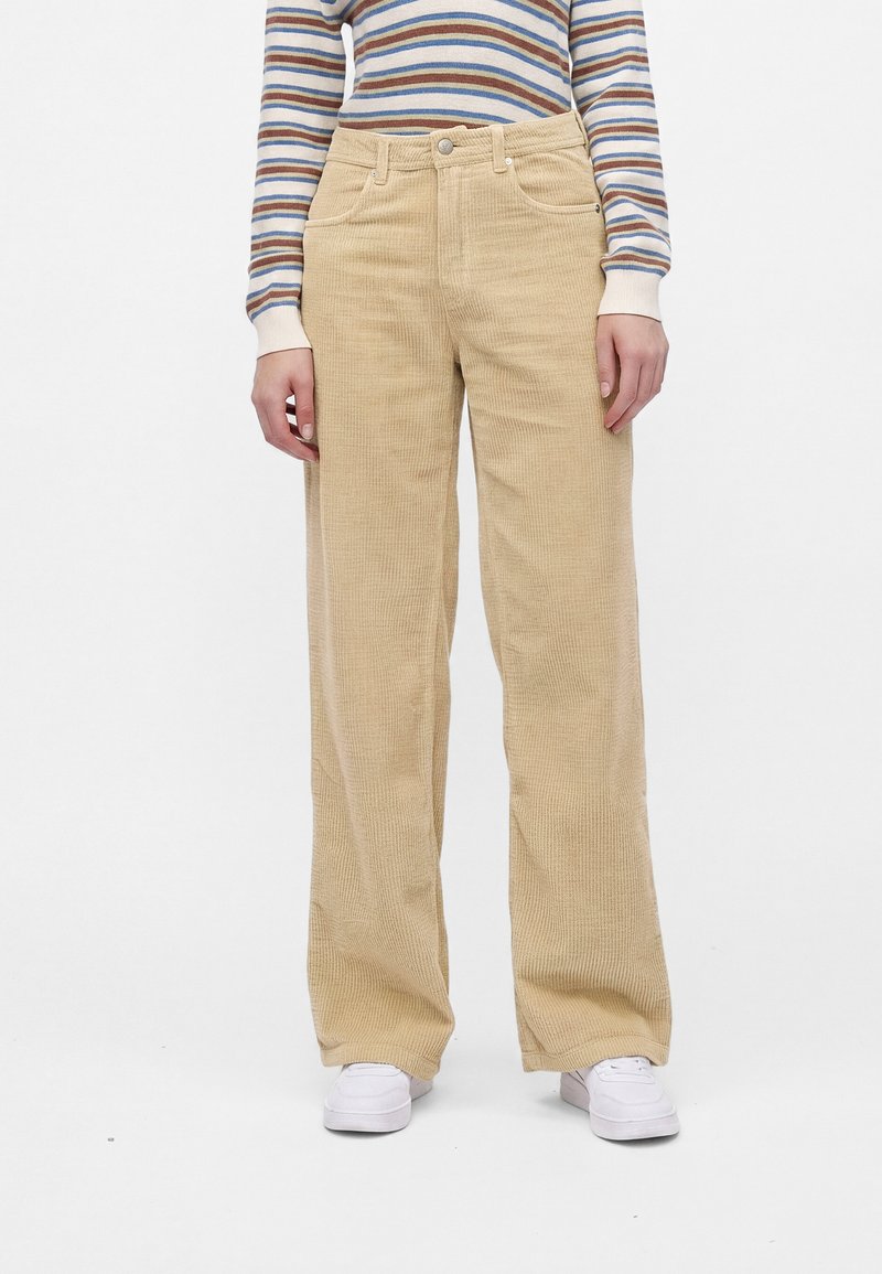 Personne portant un pantalon large en velours côtelé beige, des baskets blanches et une chemise à manches longues rayée aux couleurs bleu, marron et blanc.