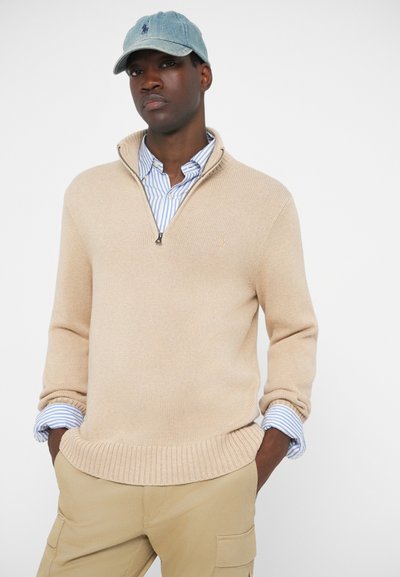 Beige kötött pulóver, negyed cipzárral, bordázott aljjal, világoskék és fehér csíkos galléros inggel párosítva. Farmer sapka logóval.