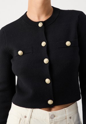 Cardigan court noir avec des boutons décoratifs dorés sur le devant et sur deux poches poitrine, porté avec un pantalon blanc.