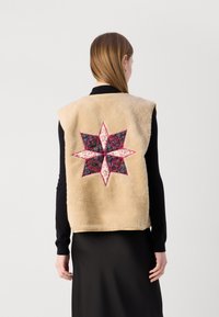 Beige fleeceväst med ett kviltat stjärnmönster i rosa och mörkt tyg på baksidan. Ärmlös design med rundad nederkant.