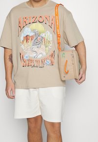Beige oversized t-shirt with a desert graphic, white shorts, and a beige bag featuring an orange strap and a green label.