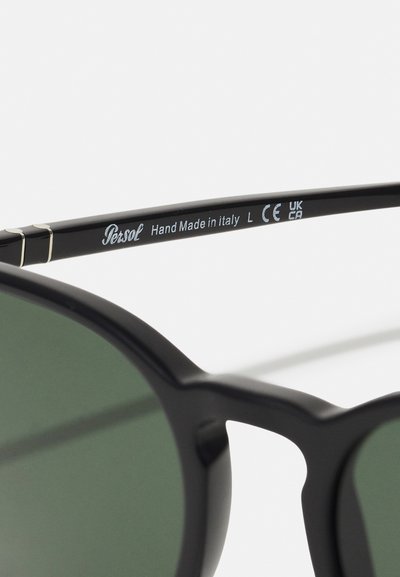 Black sunglasses with green lenses, featuring a glossy plastic frame and silver accent details on the temples. Marked 'Persol' and 'Hand Made in Italy.'