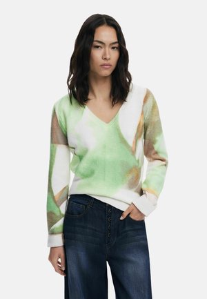 Knit sweater with a V-neckline, featuring green and beige abstract patterns. Ribbed cuffs and hem, paired with dark denim jeans.