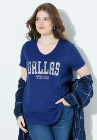 Žena s hnědými vlasy, oblečená v modrém tričku "Dallas Texas" a džínech, držící modrou kostkovanou košili, stojí na bílém pozadí.