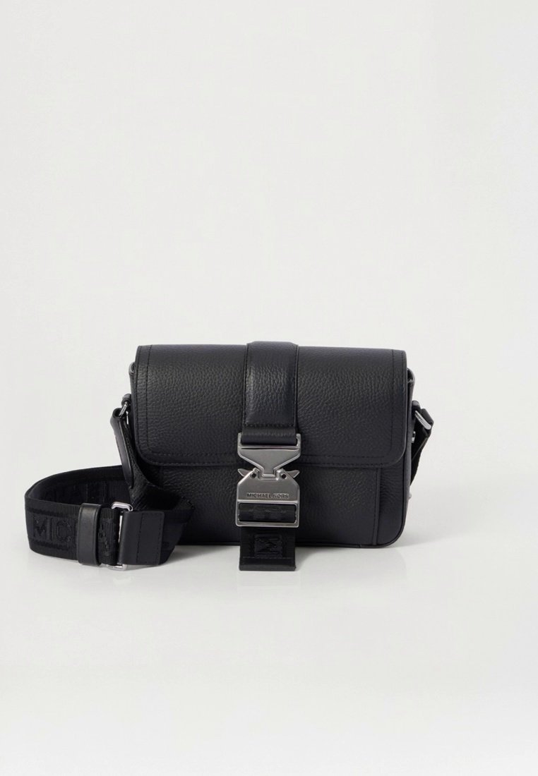 Sac bandoulière en cuir noir avec une surface texturée, une quincaillerie argentée et une bandoulière ornée d'un logo. Fermeture à pression à l'avant.