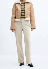 Giacca corta color tan con foderatura in shearling, abbinata a pantaloni a gamba larga color crema. Maglione a righe verde e beige sotto. Scarpe nere con punta.